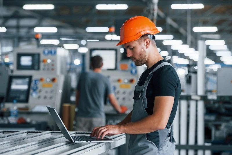 Image d'un technicien en uniforme de travail et casque de sécurité utilisant un ordinateur portable dans une usine pour planifier des opérations de maintenance.
