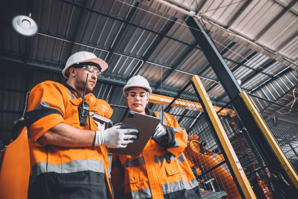 Deux ouvriers en tenue de sécurité orange consultent une tablette dans une usine, illustrant la maintenance corrective.