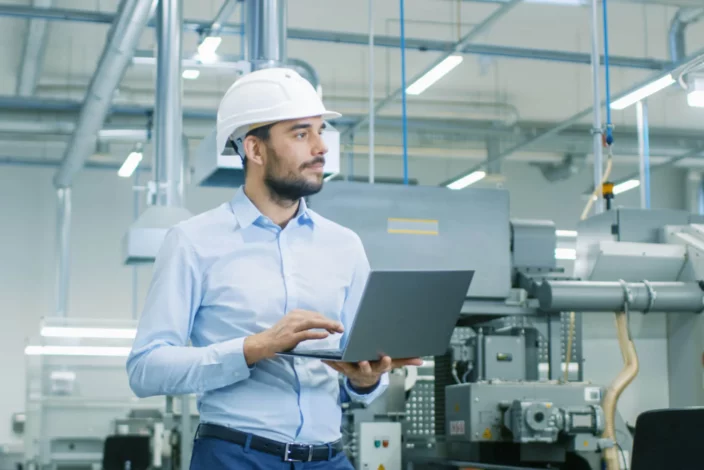 Un homme en casque de sécurité utilise un ordinateur portable dans une usine, illustrant les avantages de la maintenance corrective.