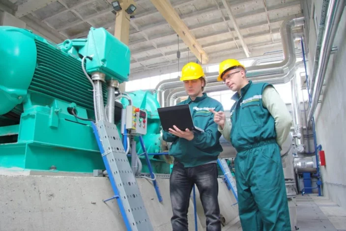 Deux techniciens en uniforme vert utilisent un ordinateur portable pour discuter de la maintenance d'une machine industrielle, illustrant l'utilisation de la roue de Deming pour l'amélioration continue.