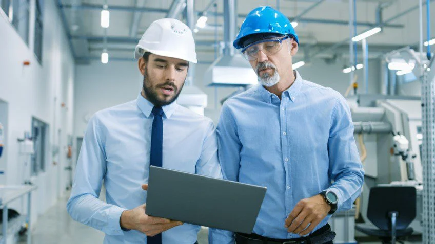 Deux hommes en casque de sécurité consultent un ordinateur portable dans une usine, illustrant l'utilisation d'un outil de maintenance corrective.